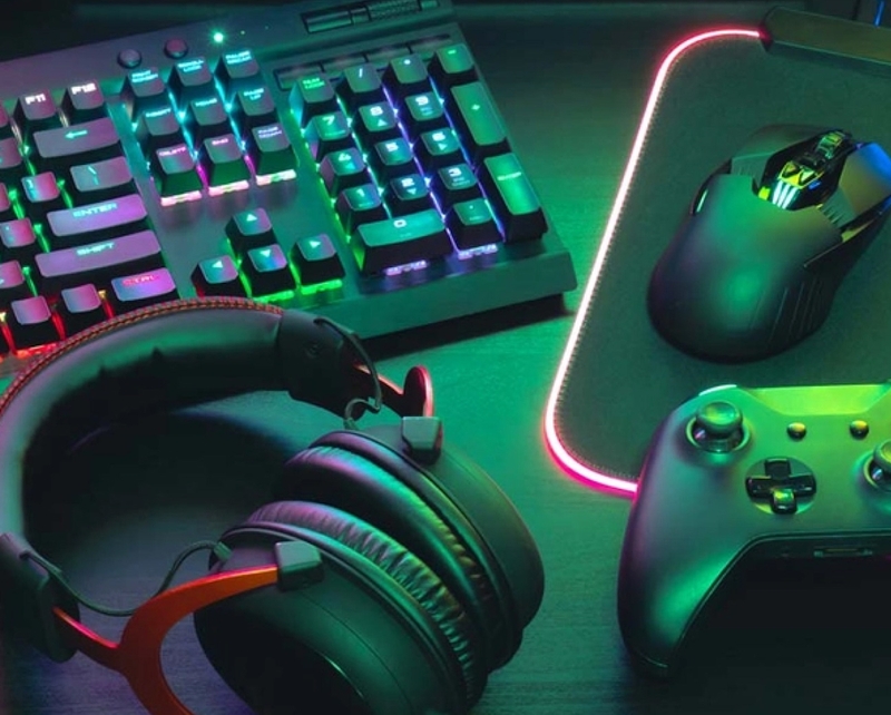 Tastatur, Kopfhörer, Maus, Handy und Spielkonsole liegen auf einem Tisch und sind in Neonfarben beleuchtet.