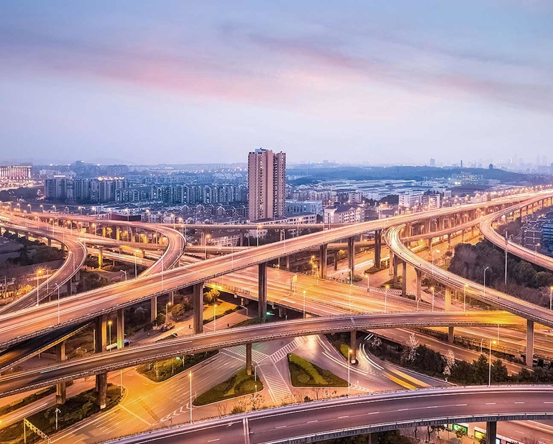 Ein riesiges, beleuchtetes Autobahnkreuz mit einer Großstadt am Horizont.